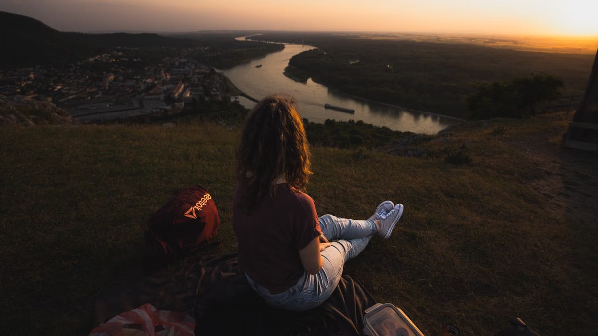 Woman sits on hillside overlooking river and town at sunset, creating a serene landscape scene.